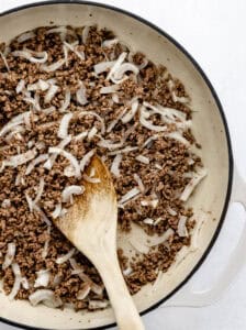 Ground beef and onion in skillet with wooden spoon.