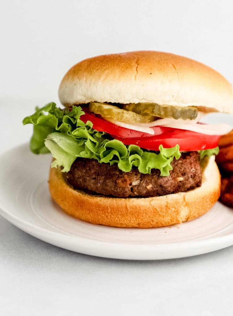 Close up front view of finished hamburger on small white plate with fries in the background.