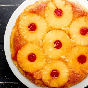 Overhead view of finished pineapple upside down cake on white plate on black tile surface.