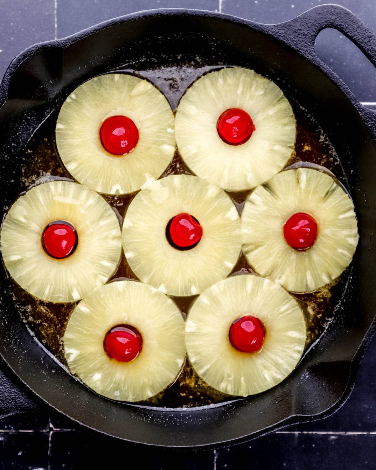 Pineapple slices and cherries added to the bottom of a cast iron skillet over brown sugar mixture. 