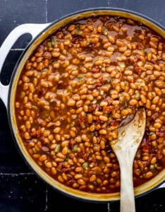 Baked bean ingredients combined in large braiser with wooden spoon.