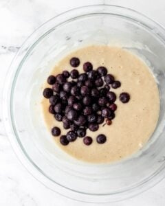 Frozen blueberries added to the other muffin ingredients in a glass mixing bowl.