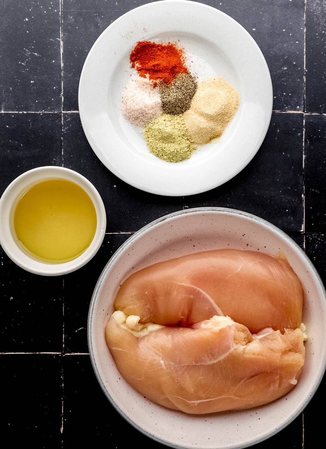 Overhead view of ingredients needed to make chicken in three separate bowls on black tile surface.
