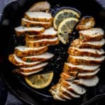 Sliced chicken and lemon slices in cast iron skillet on black tile surface.