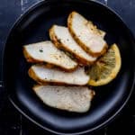 Overhead view of sliced chicken on small black plate on black tile surface.