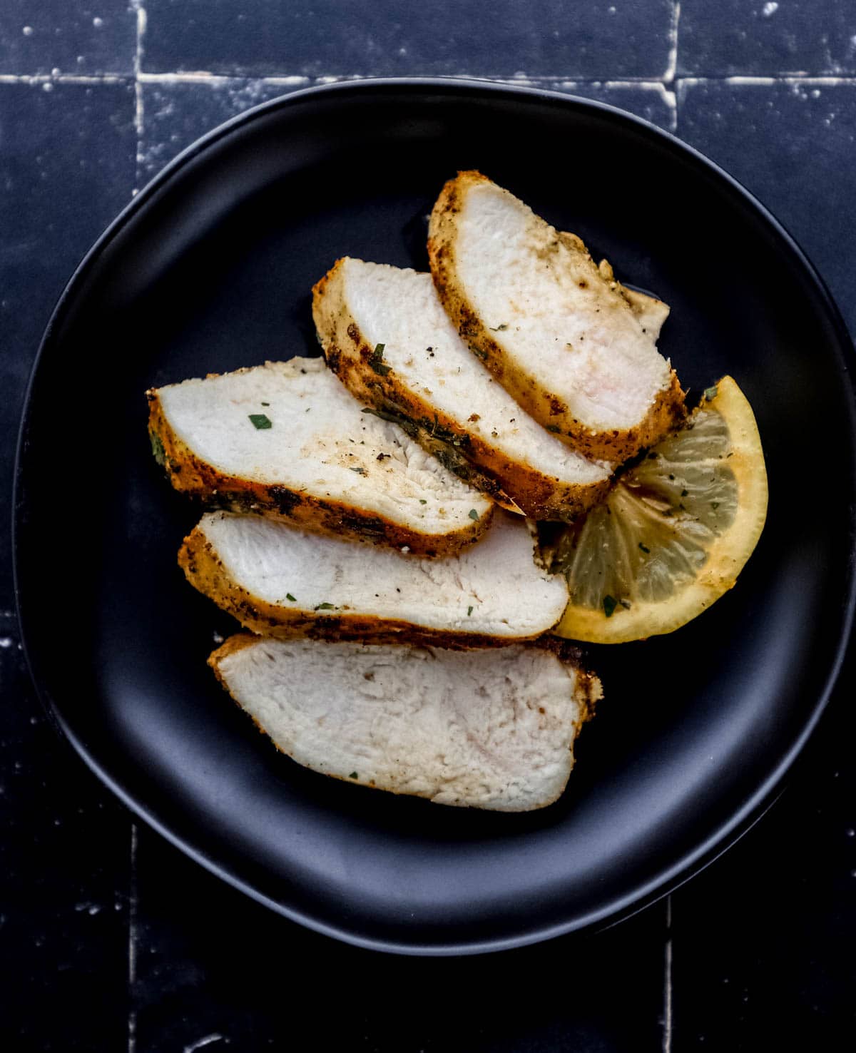Overhead view of slices of chicken and lemon on small black plate on black tile surface.