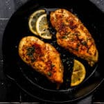 Overhead view of two pieces of cooked chicken in cast iron skillet on black tile surface.