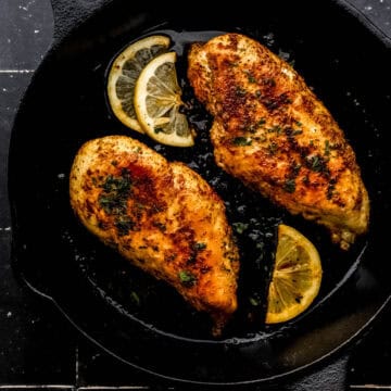 Overhead view of two pieces of cooked chicken in cast iron skillet on black tile surface.