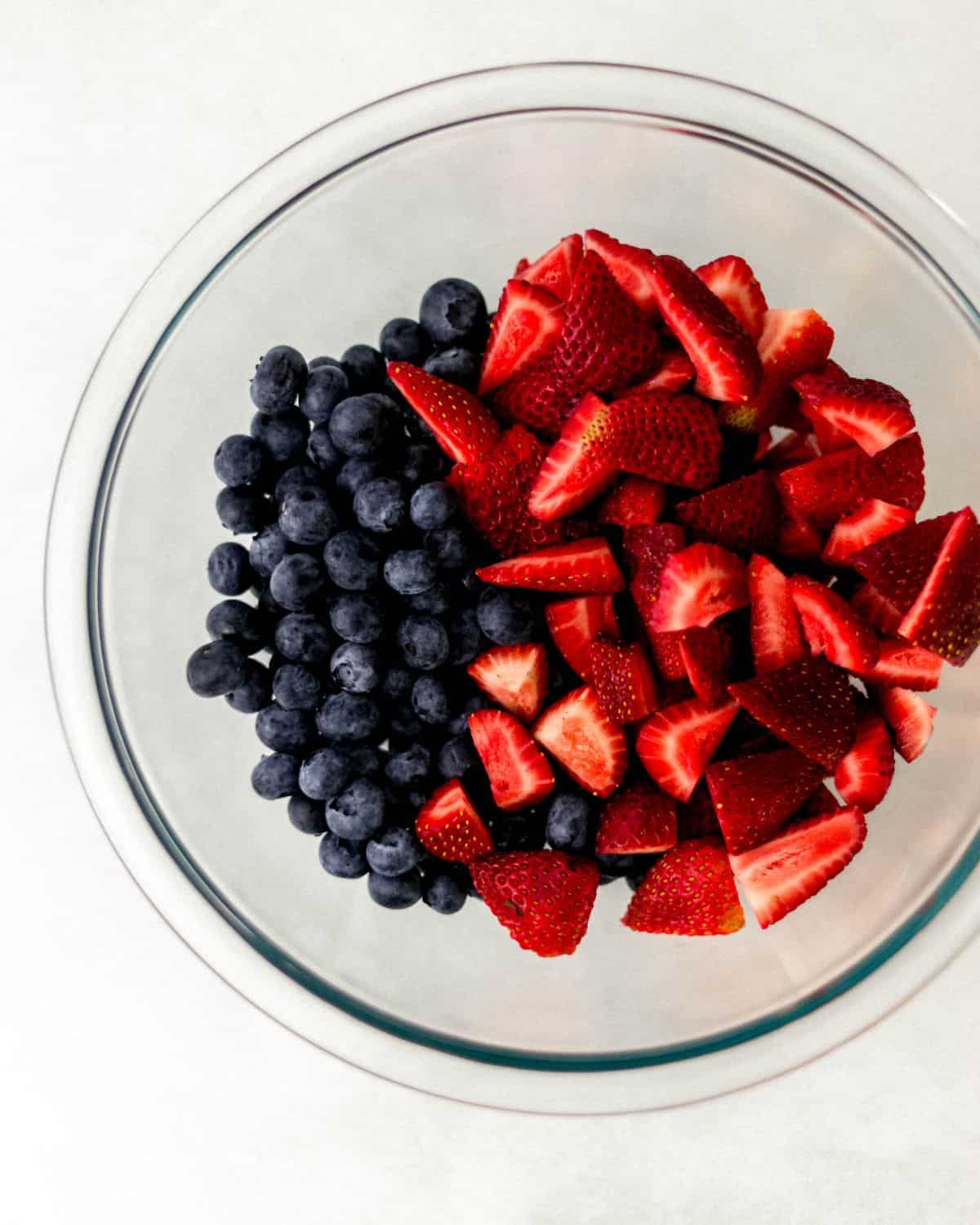 Overhead view of glass bowl with strawberries and blueberries in it on grey surface. 