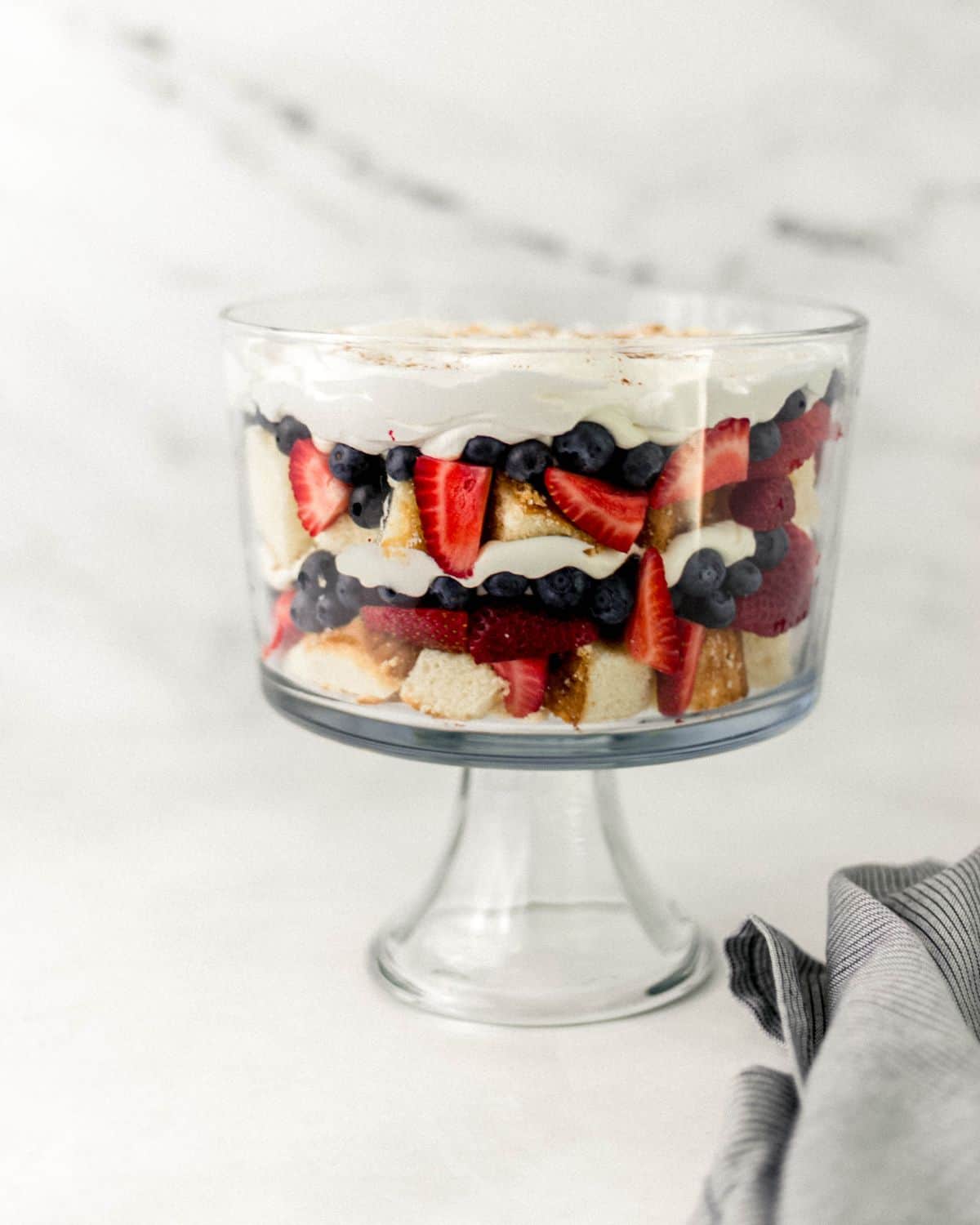 Close up side view of finished pound cake trifle in glass dish by a striped cloth napkin. 