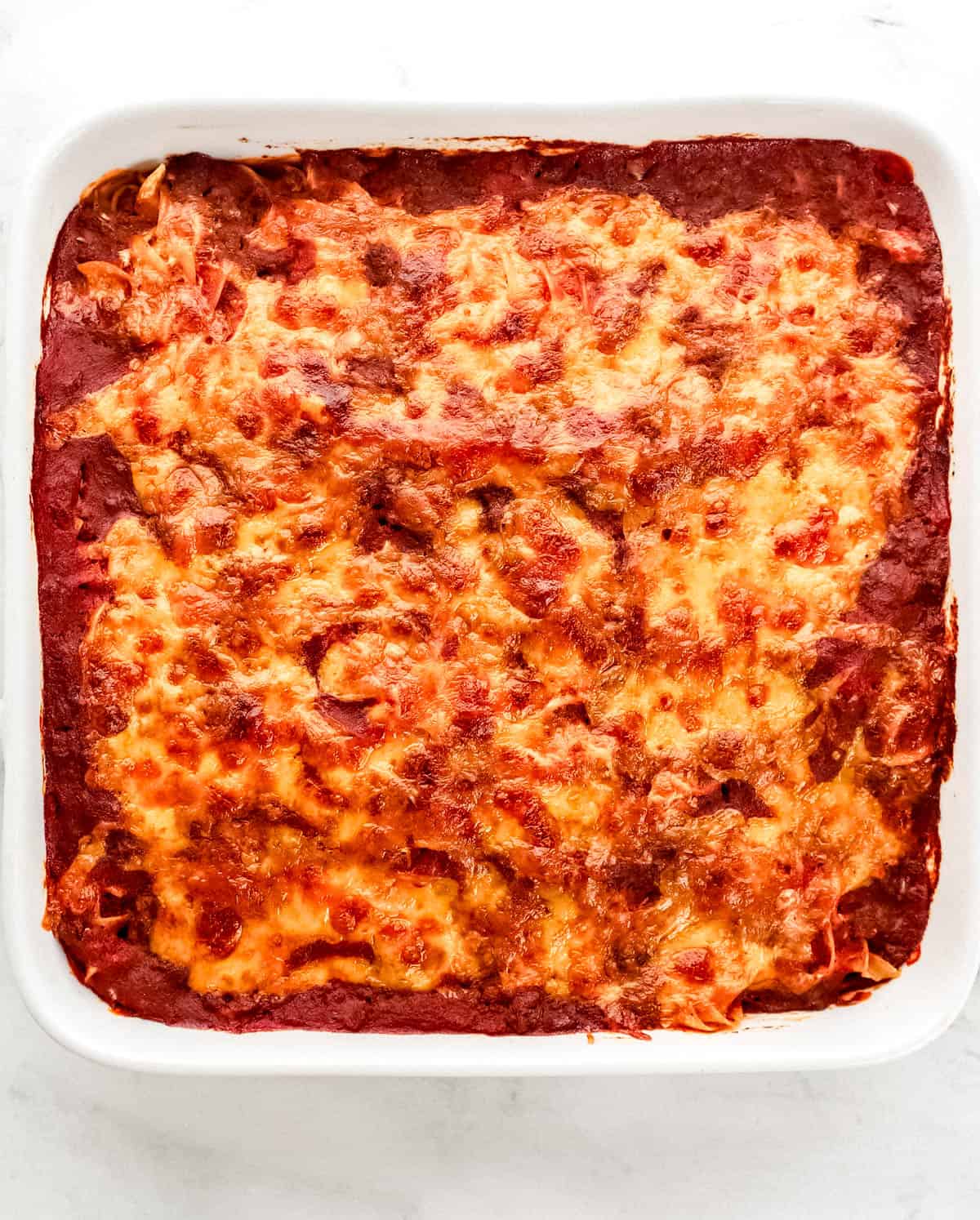 Overhead view of baked hamburger casserole in square white baking dish on marble surface. 