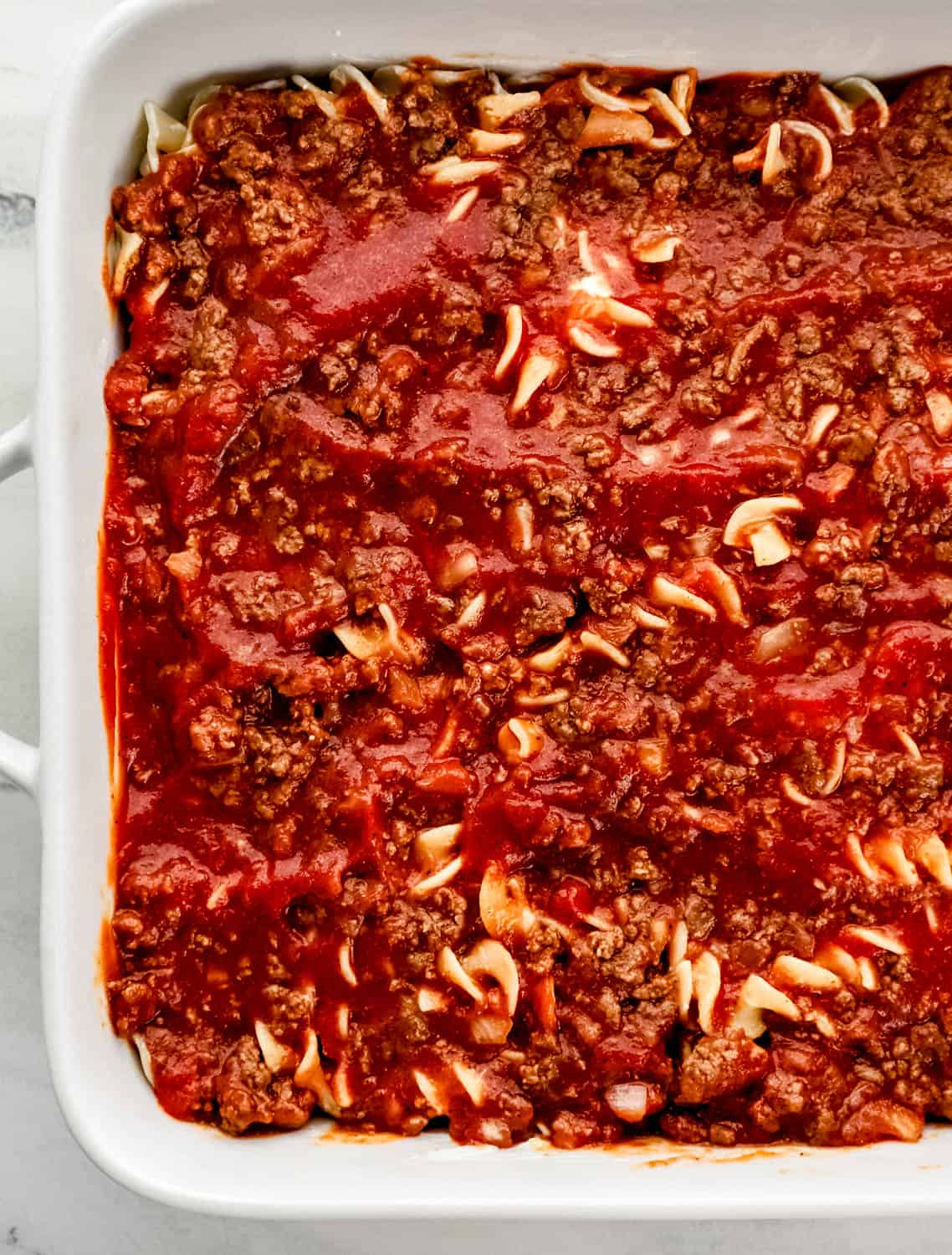 Overhead view of meat sauce added to baking dish with noodles and cream cheese mixture. 