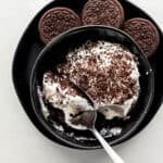 Overhead view of serving of dessert in black bowl on top of black plate with cookies on it.