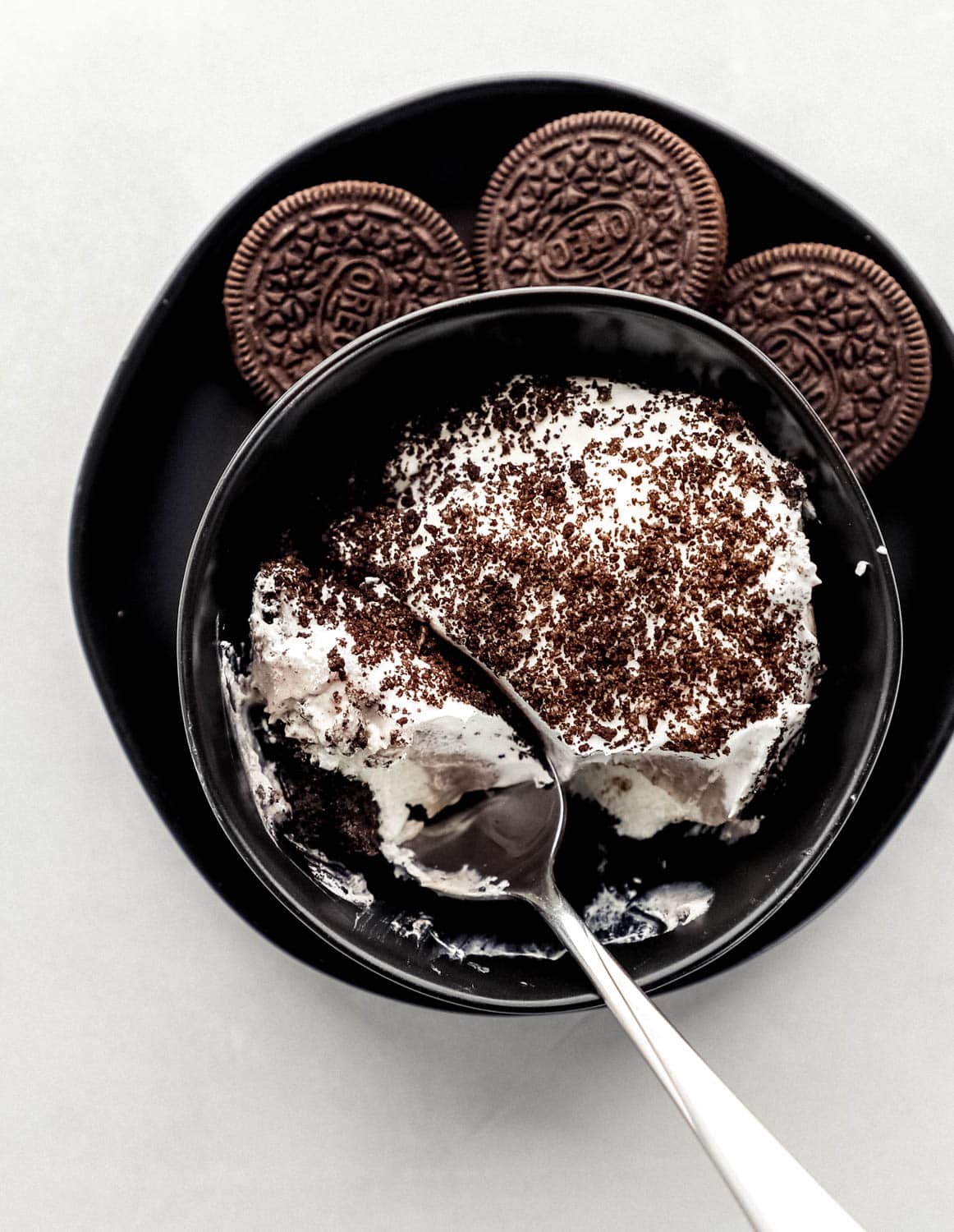 Overhead view of small black bowl with serving of dessert in it on black plate with cookies on it. 