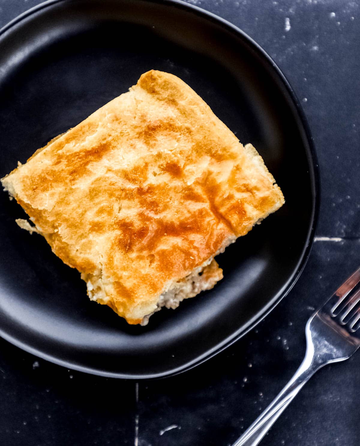Single serving of casserole on black plate by a fork on black tile surface. 