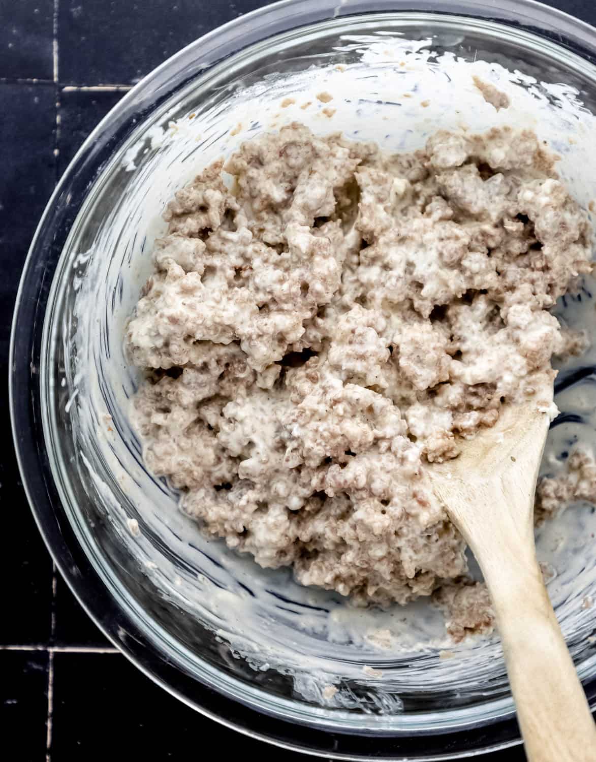 Overhead view of cooked sausage and cream cheese combined in large glass mixing bowl with wooden spoon. 