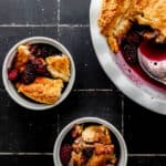 Overhead view of blackberry cobbler in baking dish and two small white bowls on black tile surface.