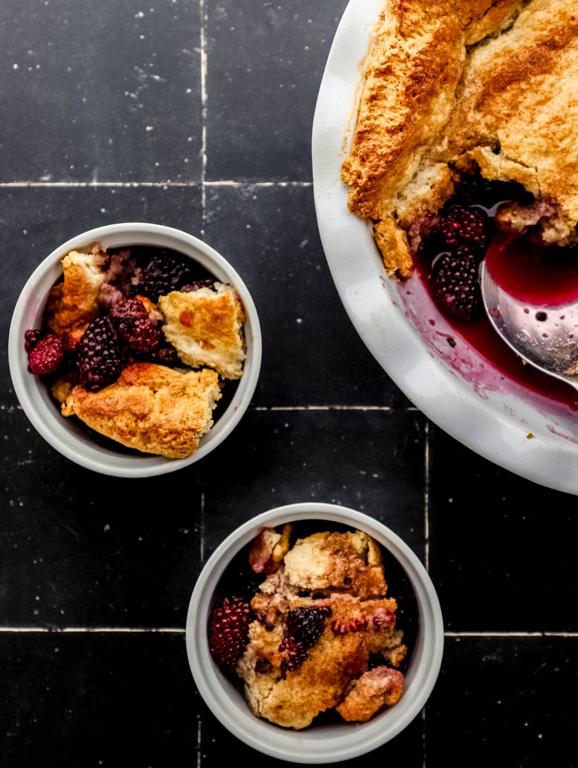 Overhead view of blackberry cobbler in white baking dish and two small white bowls on black tile surface.