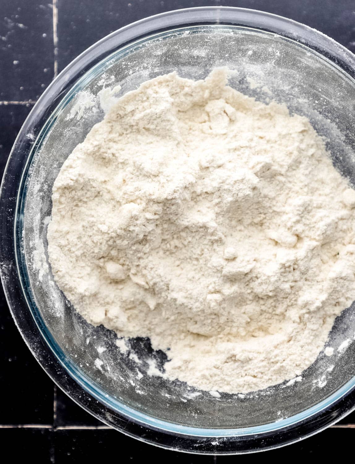 Overhead view of dry dough mixture in large glass mixing bowl on black tile surface. 