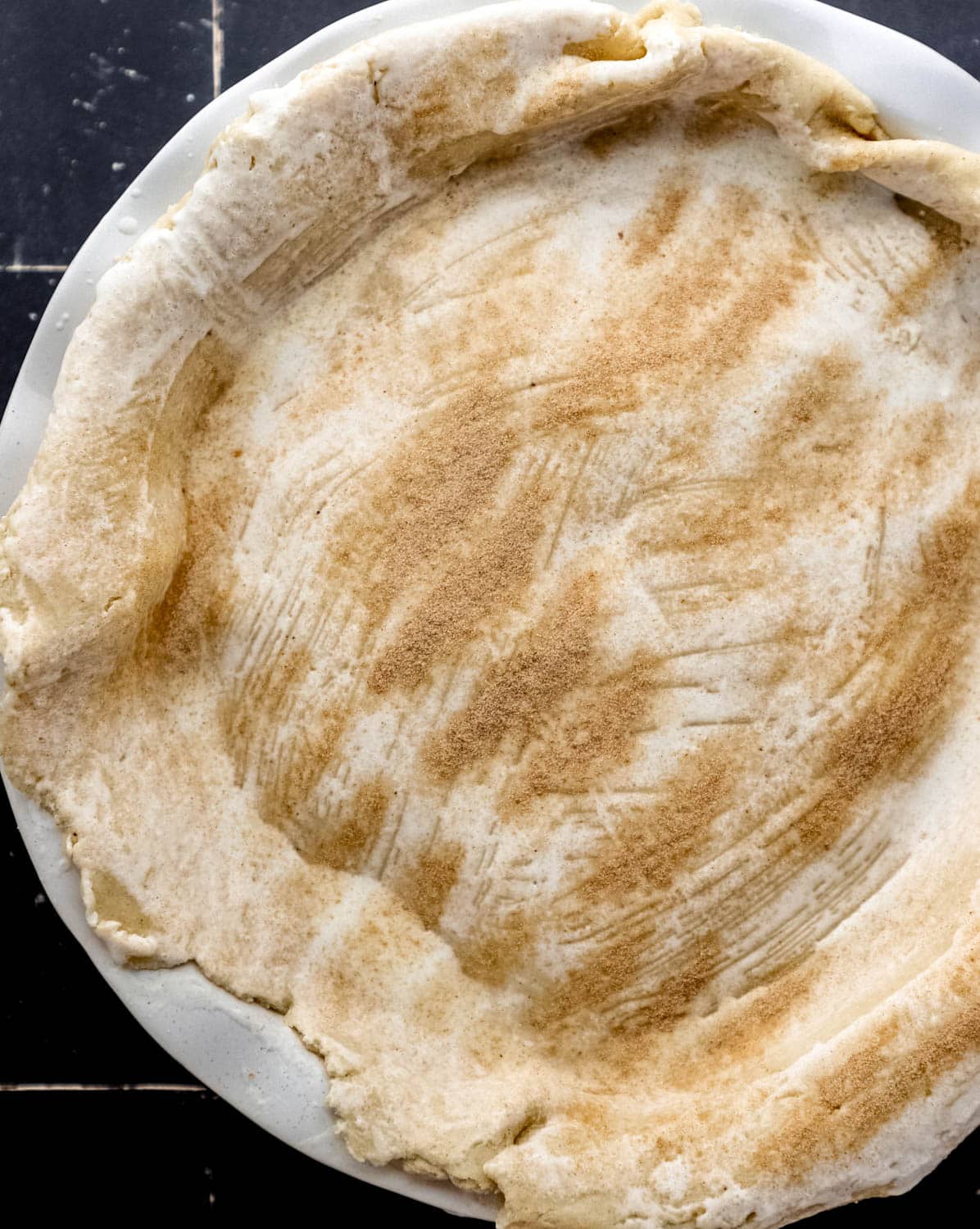 Pie crust added to blackberry cobbler filling in round white baking dish topped with cinnamon sugar. 