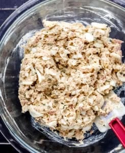 Overhead view of tuna salad ingredients combined in glass mixing bowl with spatula.
