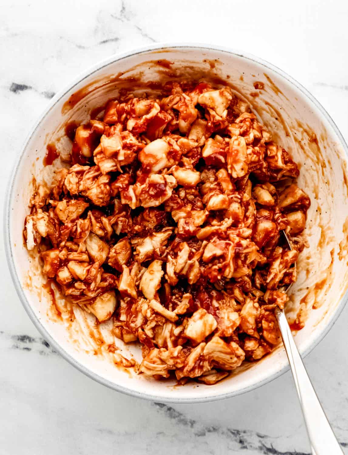 Overhead view of pulled barbecue chicken in a white bowl with a spoon in it on marble surface. 