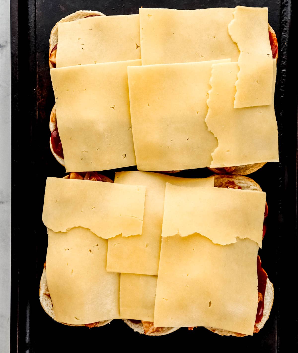 Slices of cheese added on top of bbq chicken, sauce, red onion, and buns on baking sheet. 