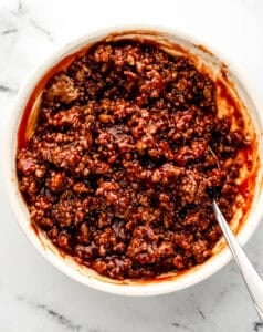 Overhead view of sauce added to ground beef in bowl with spoon.