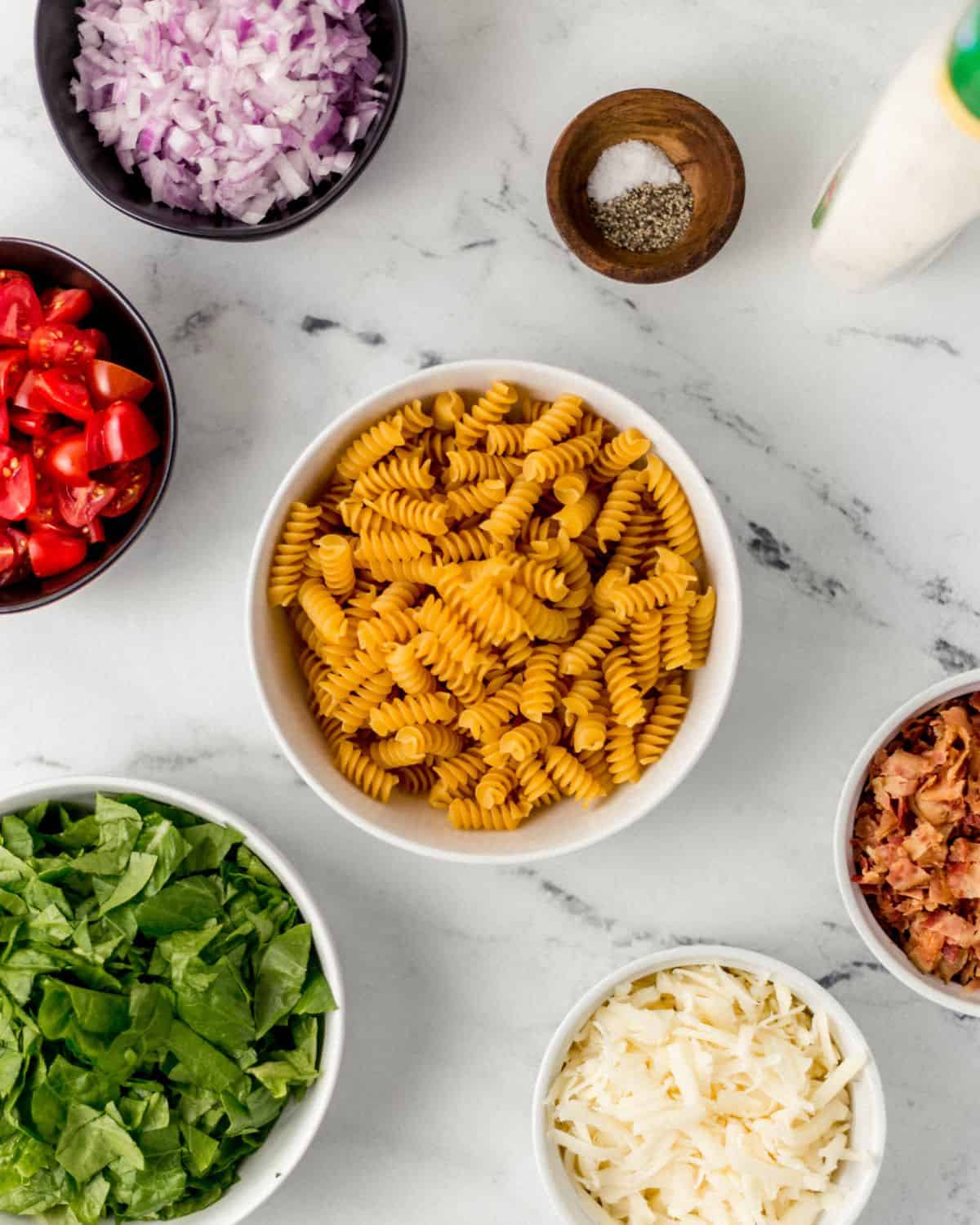 Overhead view of ingredients needed to make pasta salad in separate bowls on marble surface. 