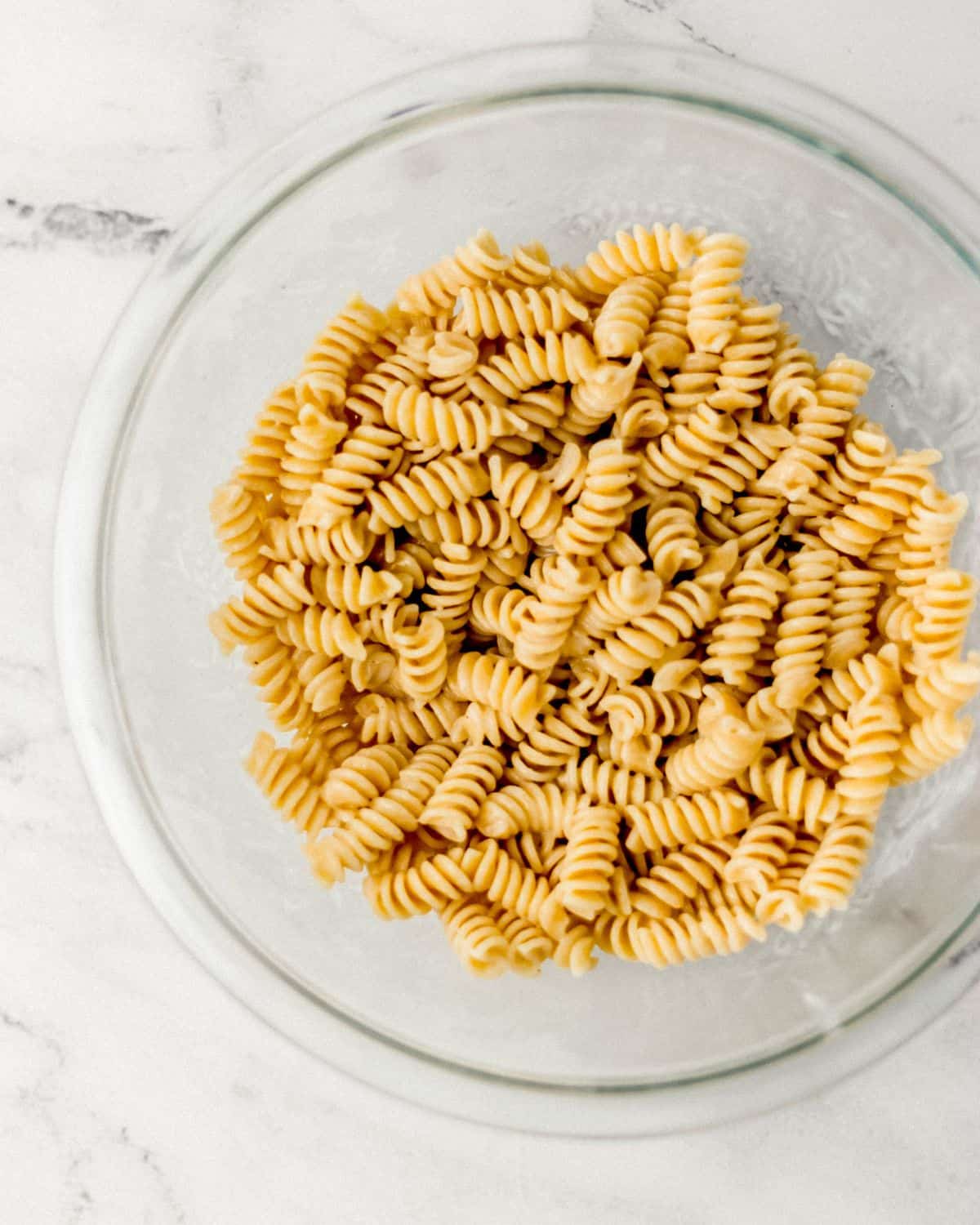 Overhead view of cooked pasta and dressing in glass mixing bowl. 