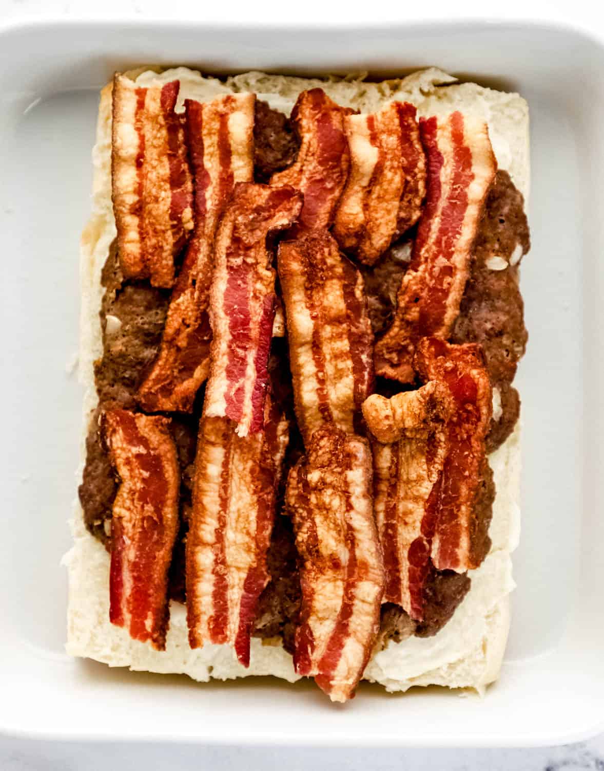 Bacon added on top of burgers on buns in white baking dish. 