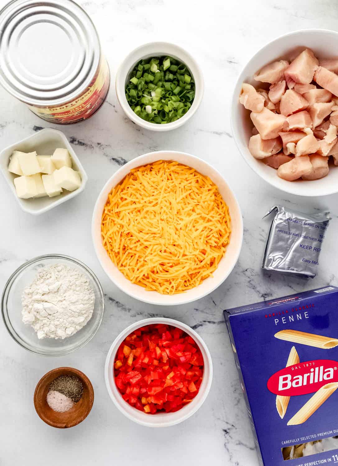 Overhead view of ingredients needed to make pasta bake in separate bowls and containers on marble surface.