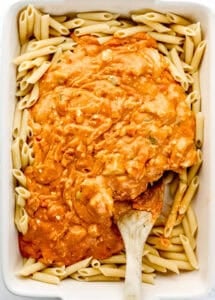 Overhead view of sauce added to pasta in baking dish with wooden spoon.