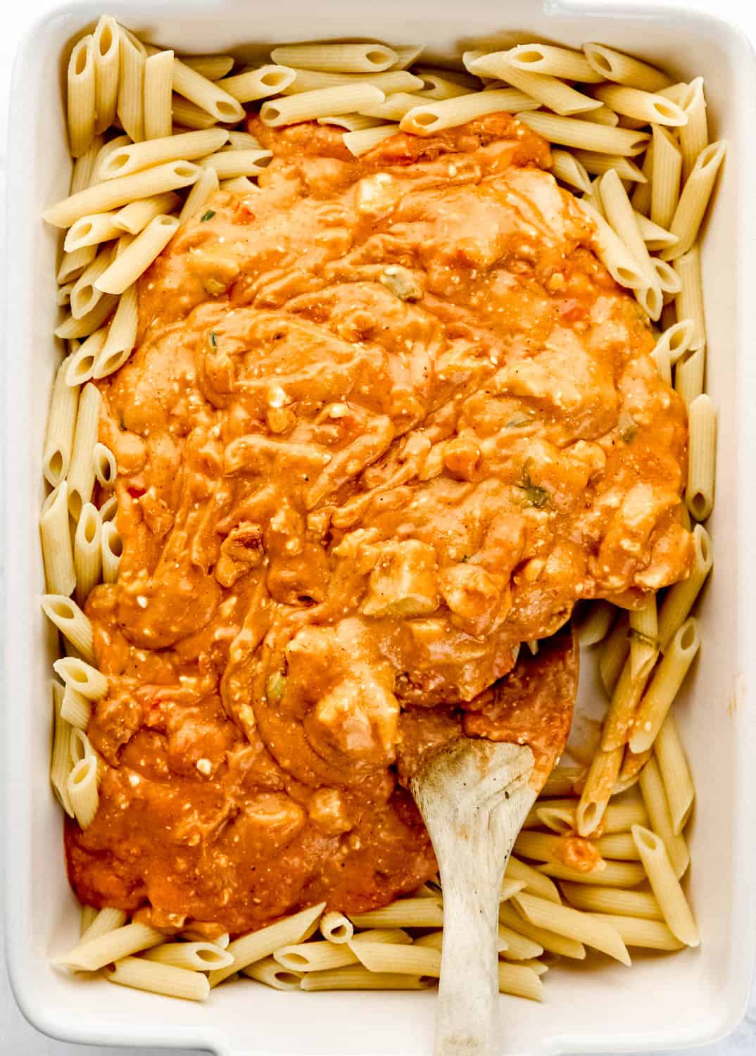 Sauce and pasta added to white rectangle casserole dish with wooden spoon in it.