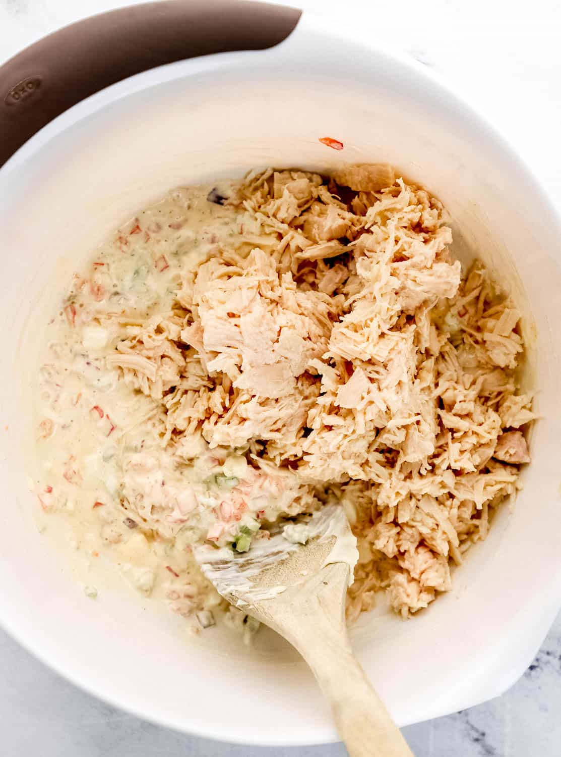 Chicken added to large white mixing bowl with the other pasta salad ingredients and wooden spoon in it. 