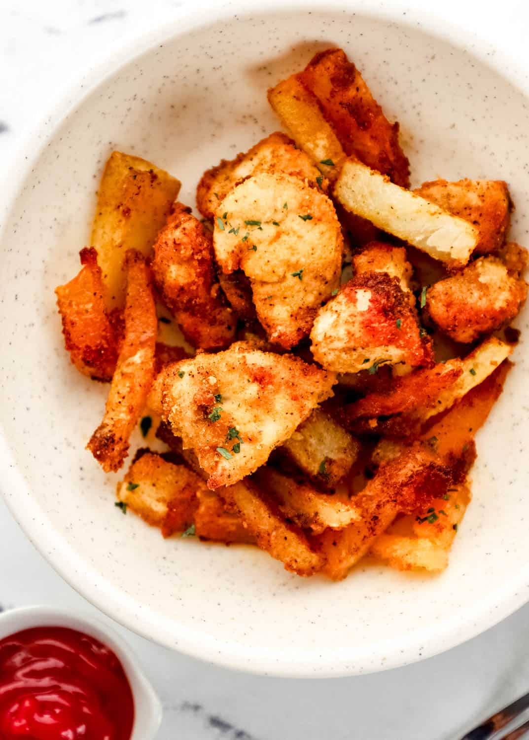 Close up overhead view of nuggets and fries in white bowl by a small bowl of ketchup. 