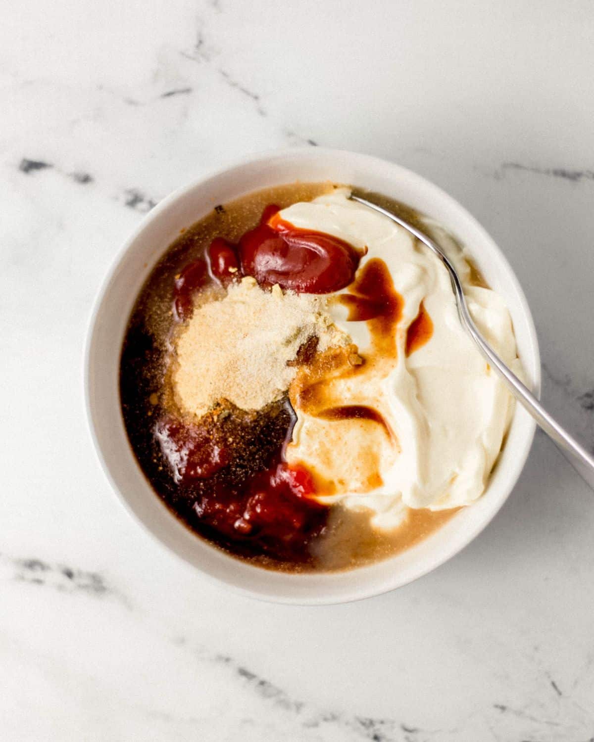 Overhead view of sauce ingredients added to white bowl with spoon in it. 