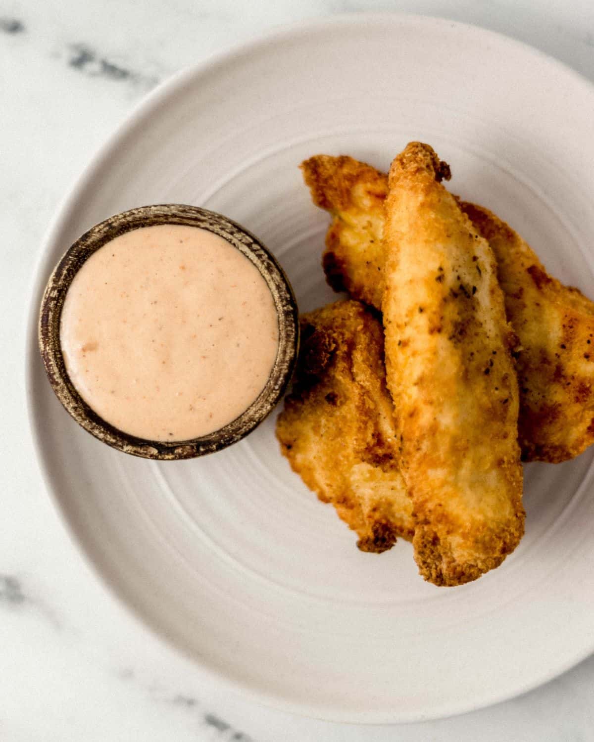 Overhead view of fried chicken tenders and small bowl of sauce in wooden bowl on plate. 