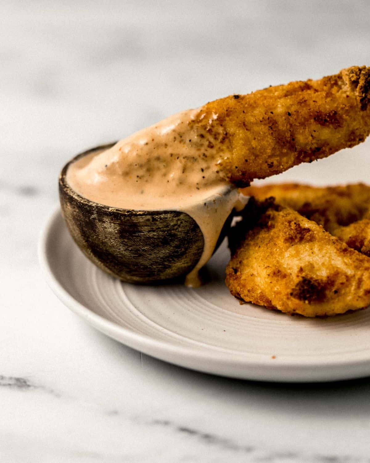 Close up side view of chicken tender being dipped in small bowl of sauce on plate. 