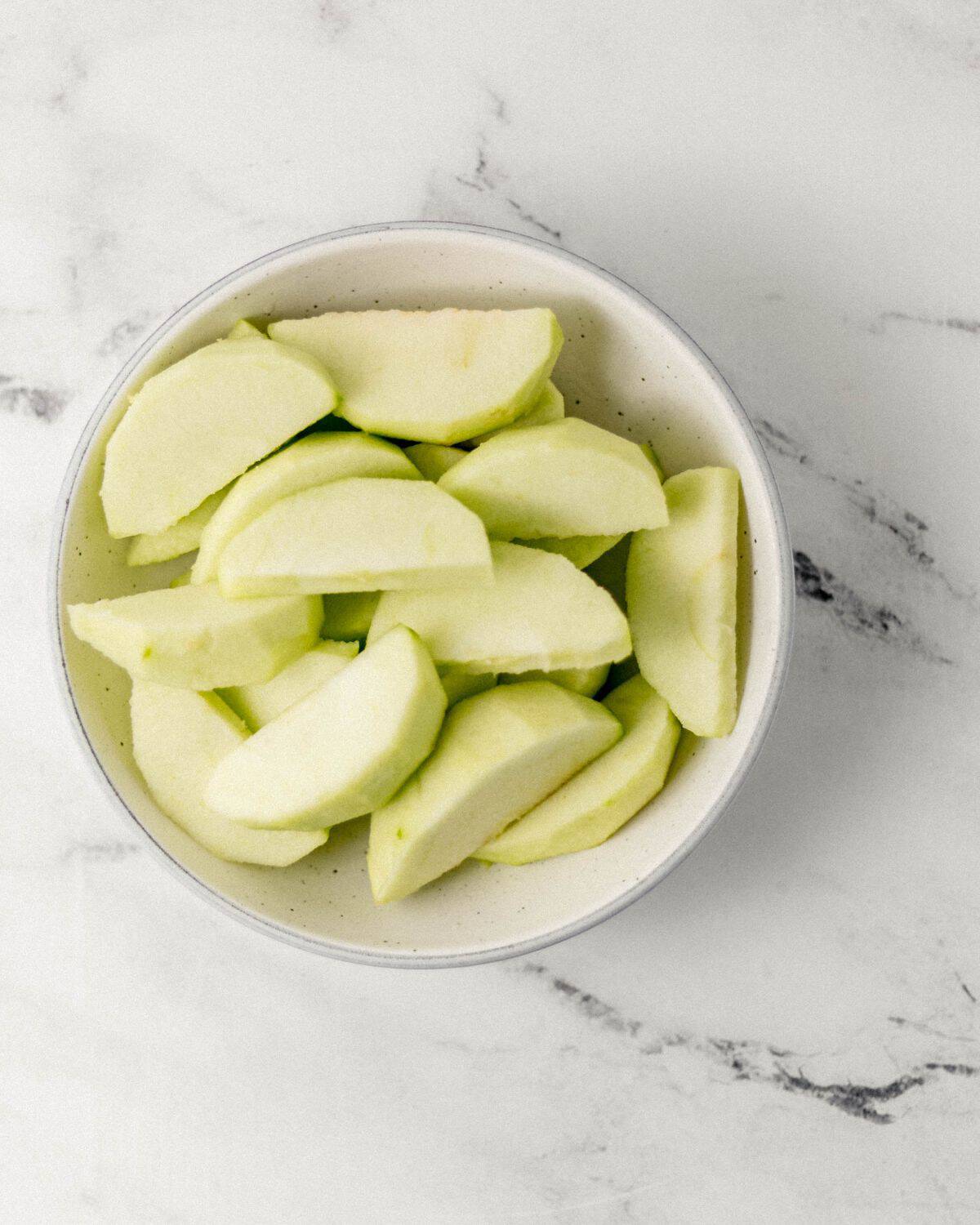 Apple slices in white bowl on marble surface. 