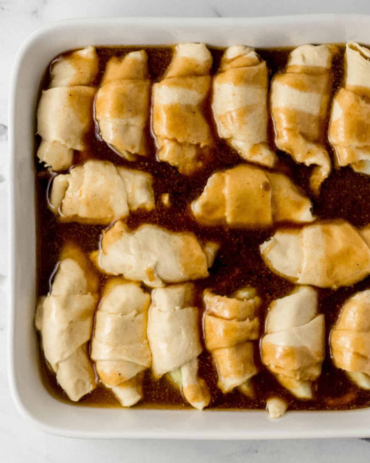 Overhead view of sauce added to crescent dough rolled apple slices in baking dish. 