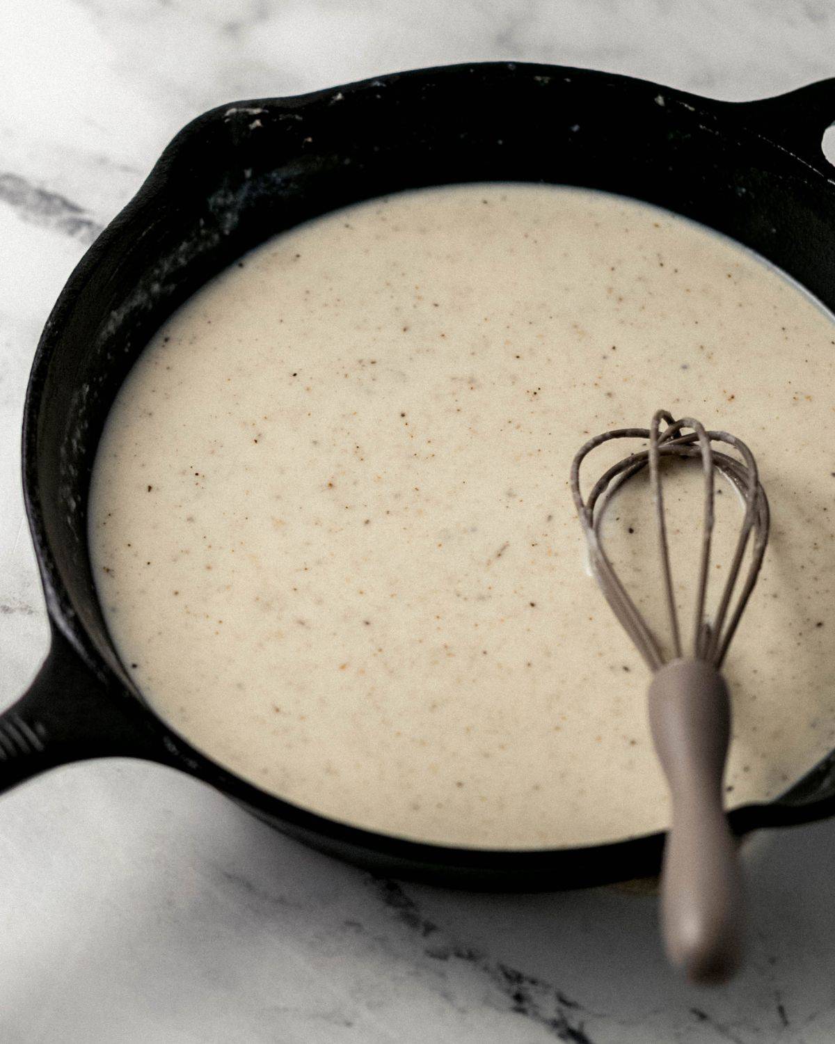 Close up side view of cast iron skillet with gravy in it with a whisk. 