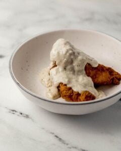Close up side view of chicken tenders topped with gravy in white bowl.