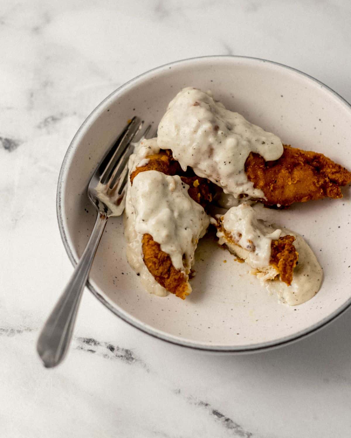 Overhead view of white bowl with fried chicken tenders topped with gravy and a fork in it. 