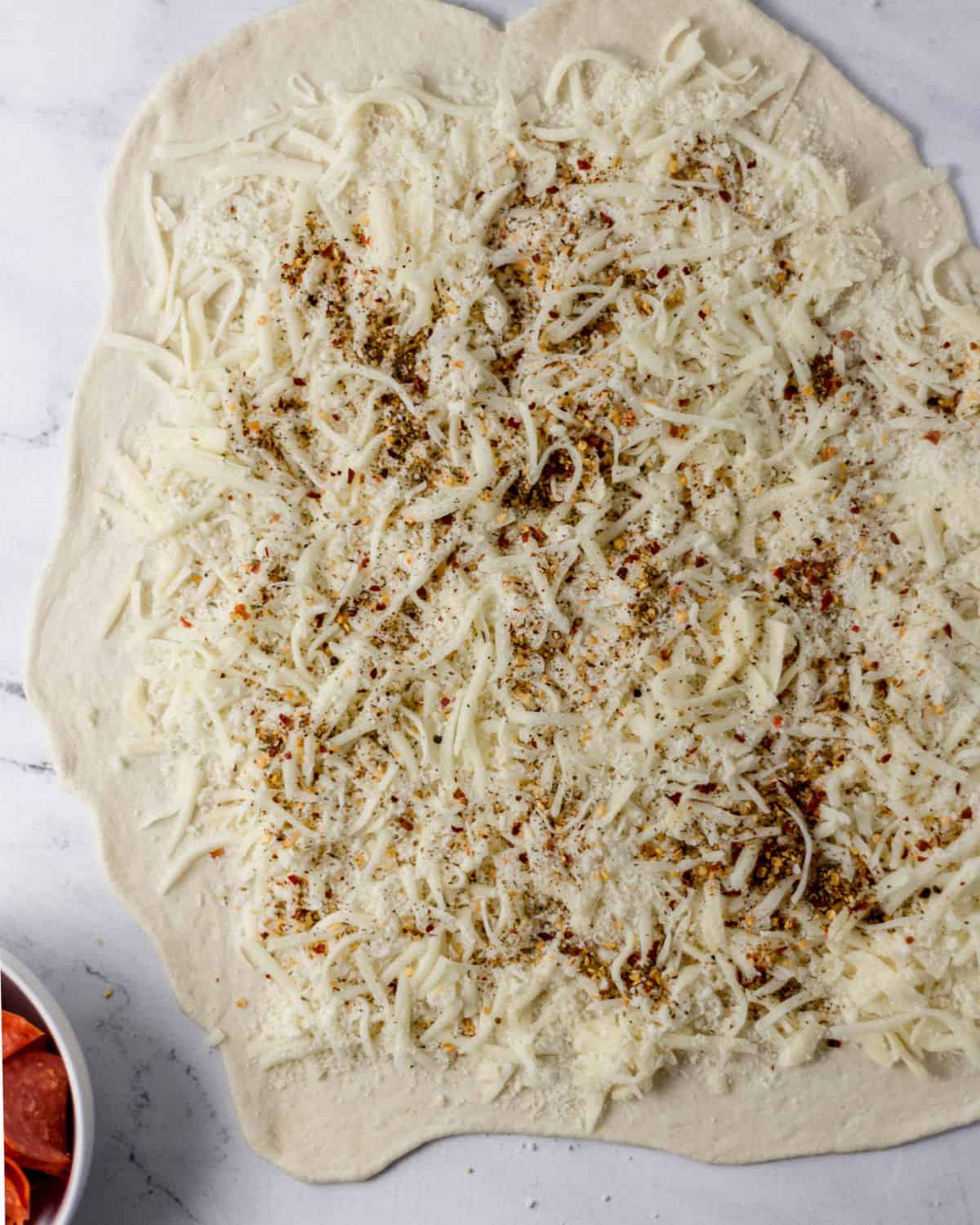 Pizza dough topped with cheese and seasoning by bowl of pepperoni. 