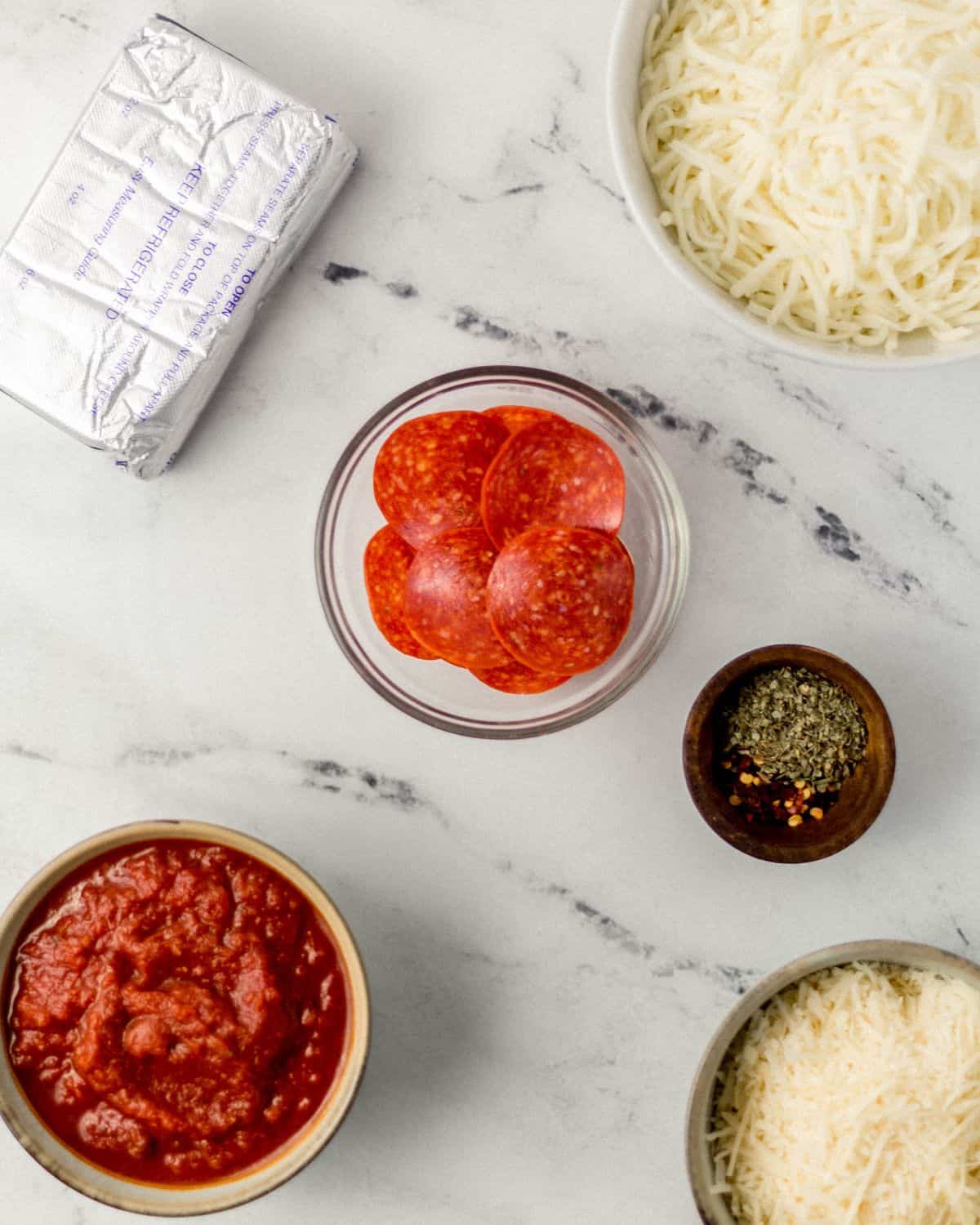 Overhead view of ingredients needed to make pizza dip in separate bowls on marble surface. 