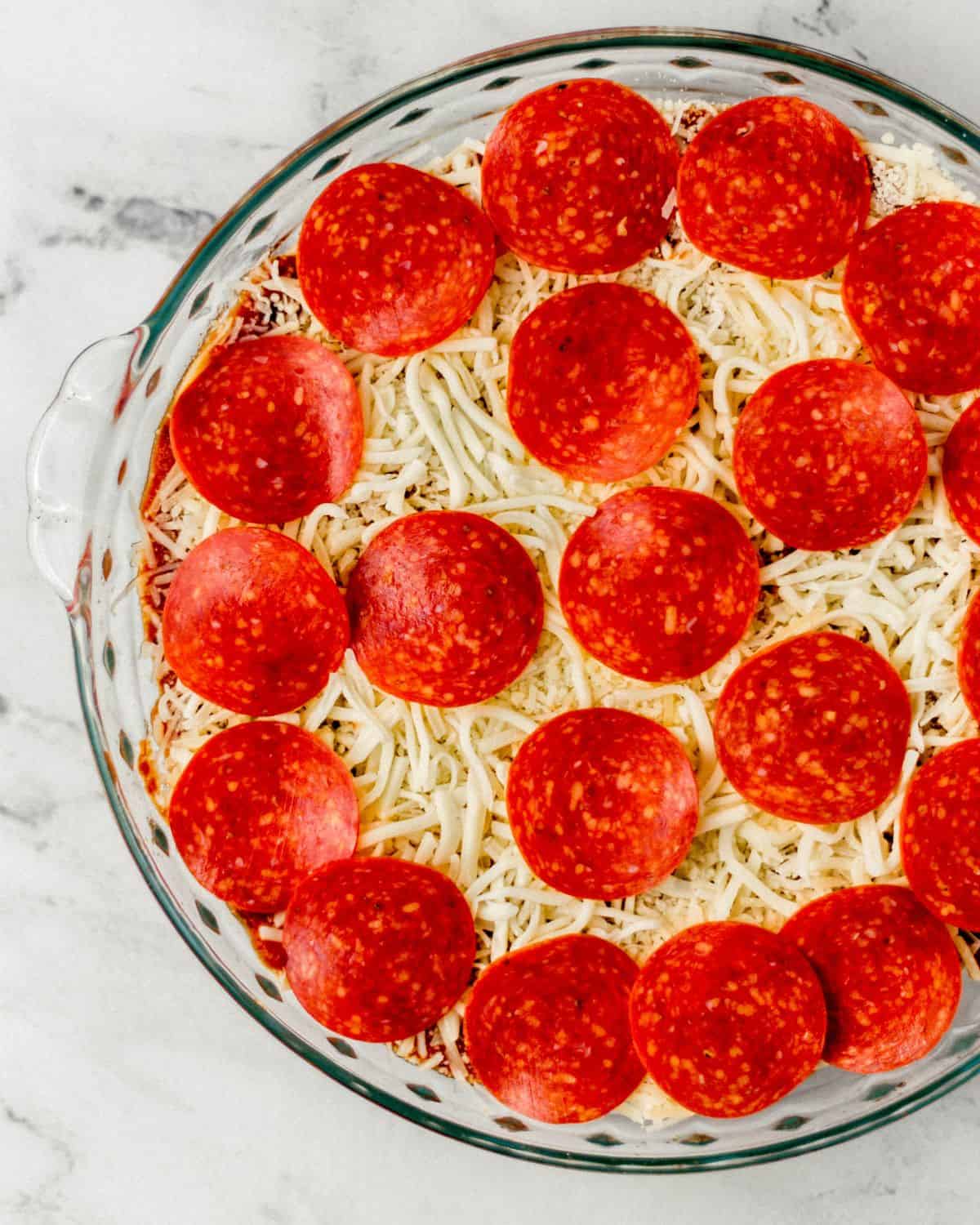 Pepperoni slices spread on top of cheese layer of pizza dip in glass baking dish. 