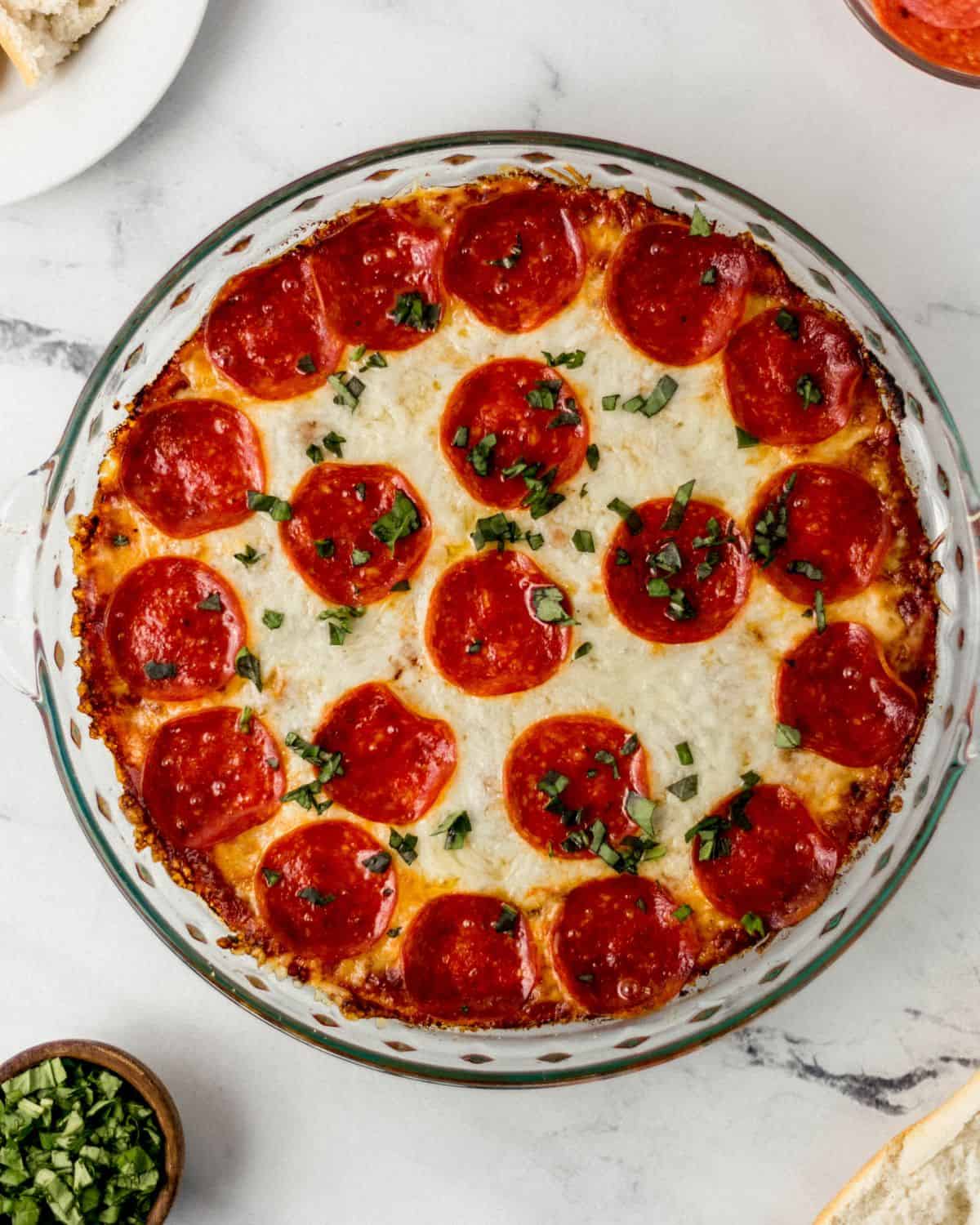 Overhead view of finished pepperoni dip in glass dish on marble surface topped with chopped basil.