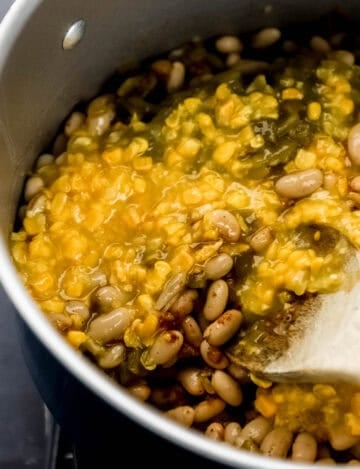 Close up side view of corn and beans added to stock pot with wooden spoon.