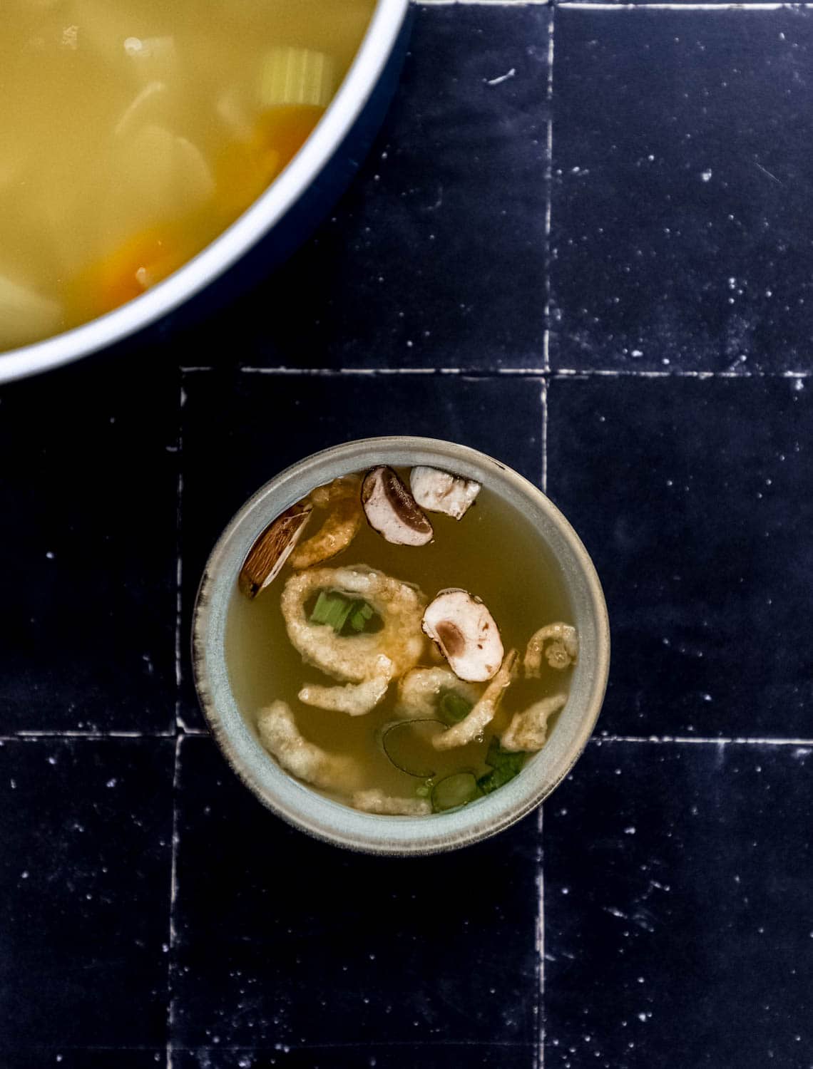 Overhead view of small bowl of soup on black tile surface with the pot of soup in the background.